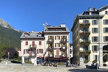 Lively European town square with people dining outside cafes, historic buildings, and majestic mountains under a clear blue sky.