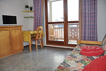 A cozy room features a TV, dining table, and a sofa with a colorful cover. A glass door opens to a balcony with carved wooden railings.