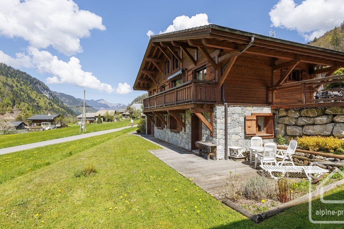 Sunny wooden and stone chalet with a balcony and outdoor patio, set in a lush green mountain valley under a clear blue sky.