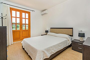 A simple, bright bedroom with a double bed, dark furniture, white walls, and light-tiled floor, featuring wooden French doors leading to an outdoor view.