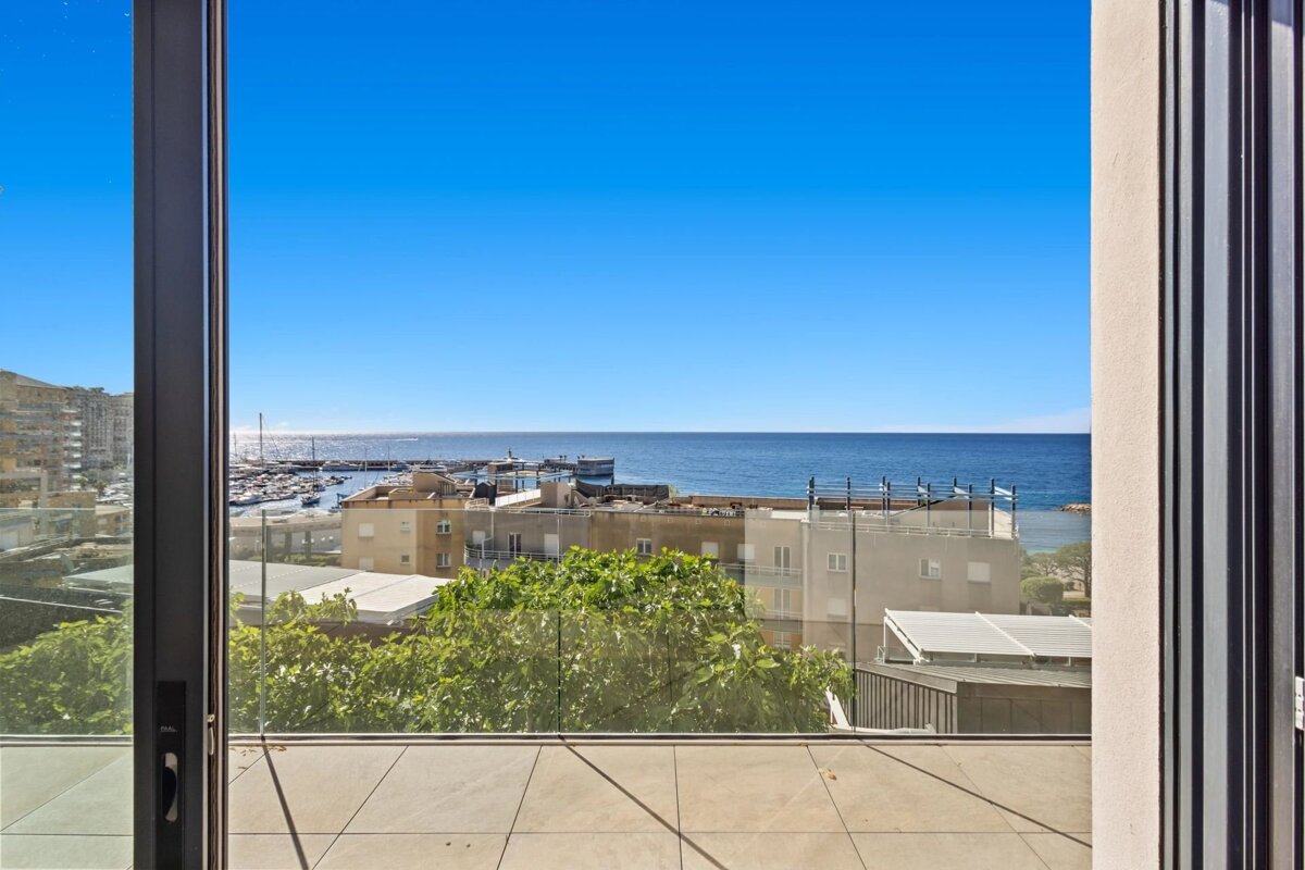 A sunny balcony view looking out over green trees, coastal buildings, and the deep blue sea under a clear sky.