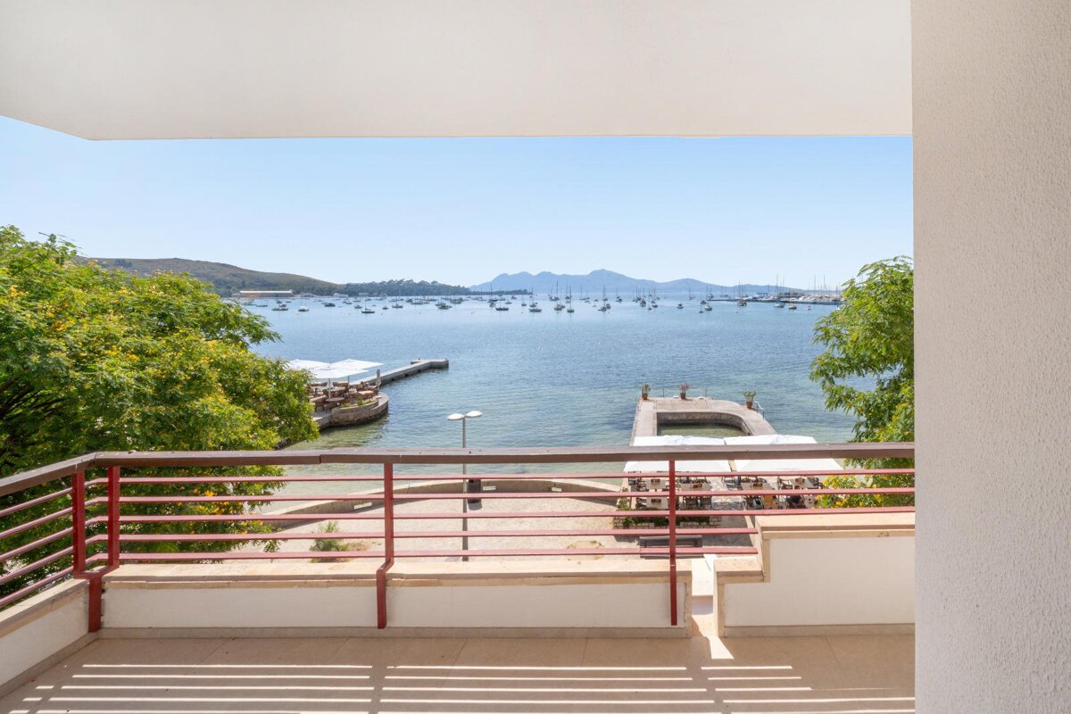 Sunny balcony view over a bay filled with numerous boats, two piers, and distant green hills under a clear blue sky. A red railing frames the scene.
