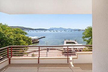 Sunny balcony view over a bay filled with numerous boats, two piers, and distant green hills under a clear blue sky. A red railing frames the scene.