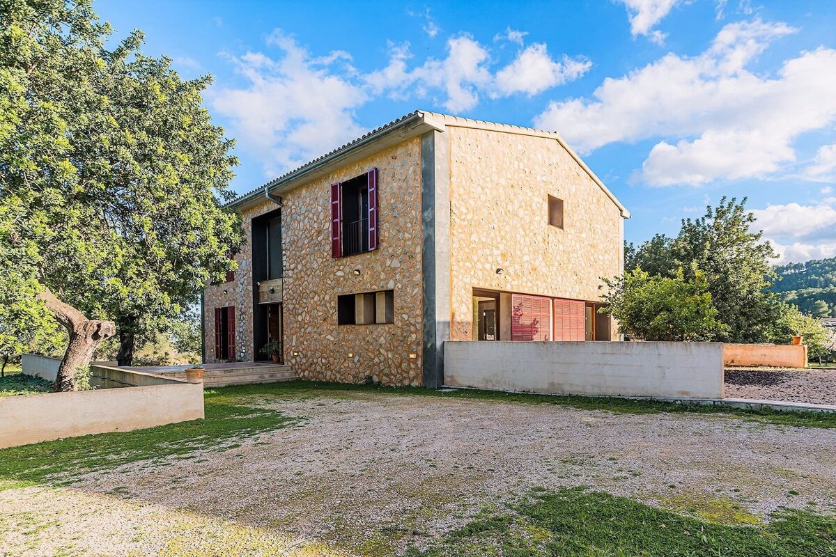 A rustic stone house with red shutters stands by a large tree, featuring a gravel yard under a bright blue sky.