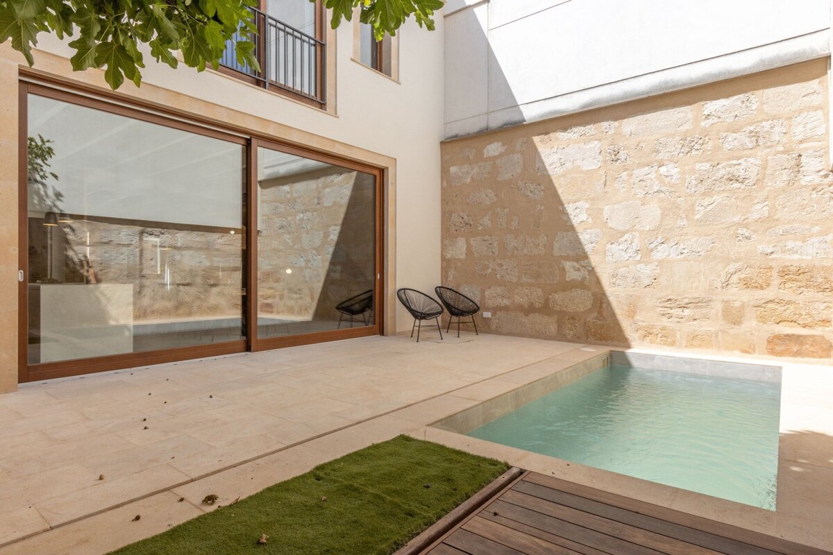 A modern patio features a small pool, large glass sliding doors, an old stone wall, and two black chairs, with sunlight creating shadows.