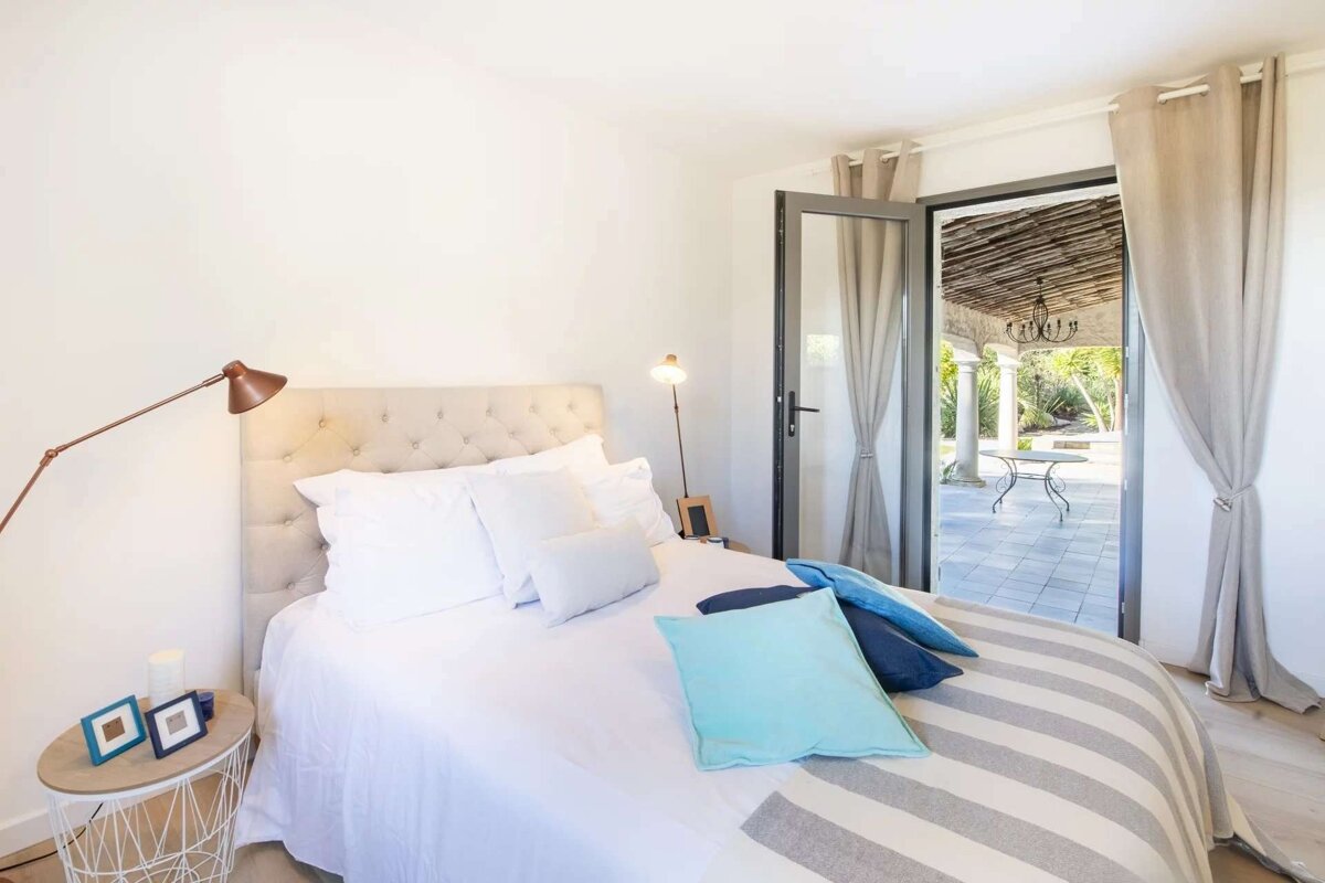 A bright white bedroom with a tufted bed, blue pillows, and open French doors leading to a beautiful outdoor patio with a chandelier and garden.