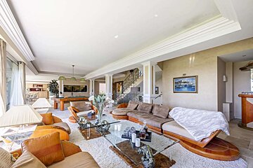 A spacious, luxurious living room featuring warm-toned sofas, a white shag rug, glass coffee tables, a pool table, and a grand staircase.