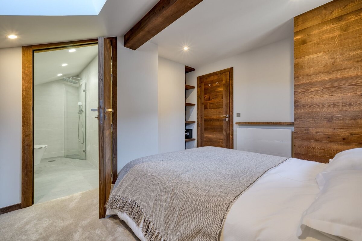 A modern bedroom features a bed with a rustic wooden headboard, white walls, and an open doorway to an en-suite bathroom with a shower.