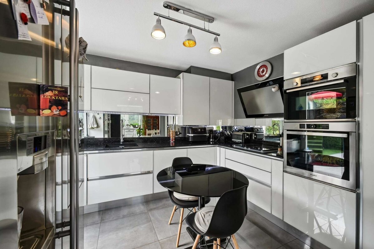 A sleek, modern kitchen featuring white glossy cabinets, black countertops, stainless steel appliances, a small black dining table, and grey tiled floors.