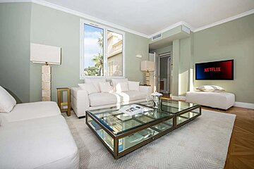 Bright, modern living room with sage walls, white sofas, glass coffee table, and a TV showing Netflix. Large window overlooks palm trees.