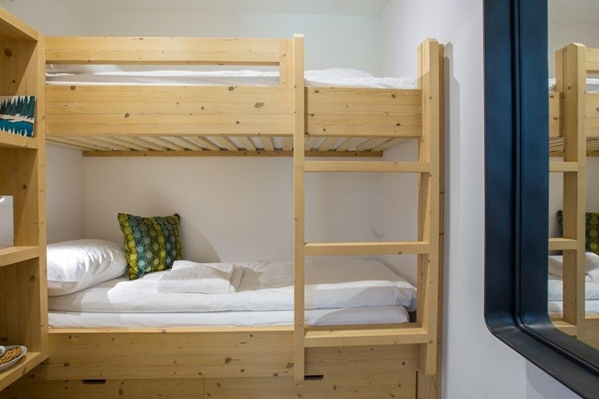 Light wood bunk beds with white bedding, a green patterned pillow, and a built-in ladder, shown next to a mirror.