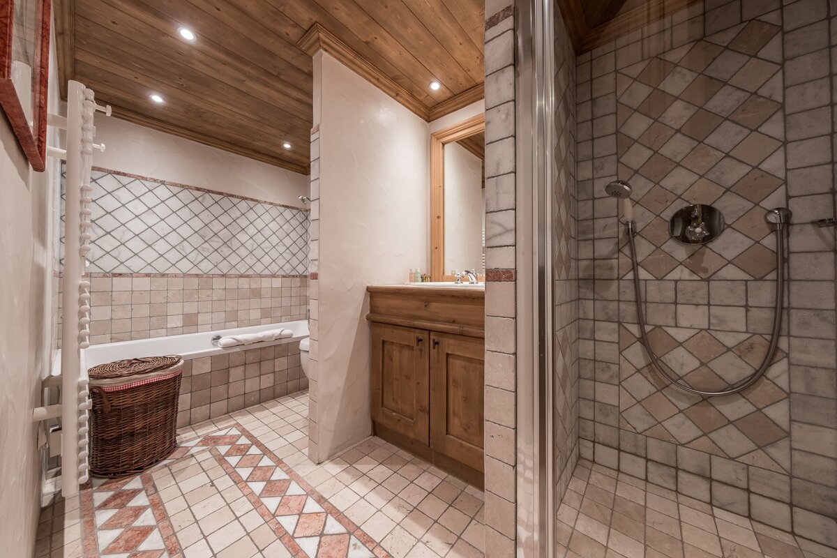 A cozy bathroom featuring patterned beige and red-brown tiles, a warm wooden ceiling, separate glass shower, bathtub, and wooden vanity.