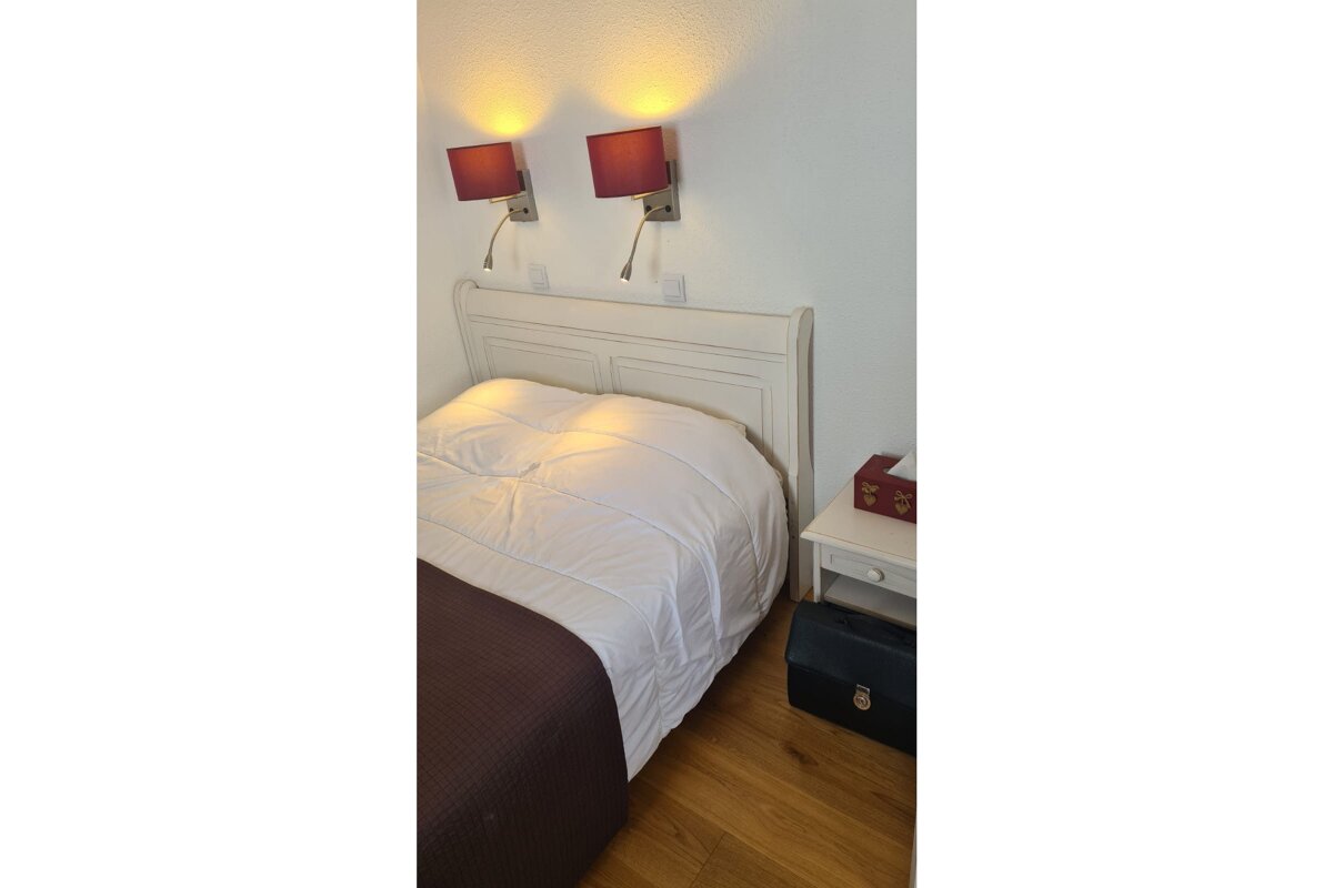 A cozy bedroom features a white bed with a headboard, two red-shaded wall lamps, and a nightstand with a tissue box on a wooden floor.