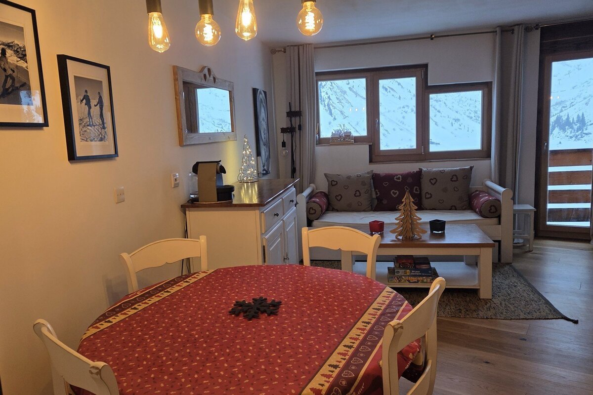 Cozy ski chalet living room with a dining table, sofa, warm lighting, and snowy mountain views outside the windows, decorated for winter.