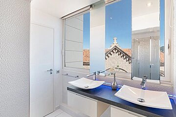 A modern minimalist bathroom featuring dual white vessel sinks and a large window with views of rooftops and blue sky.
