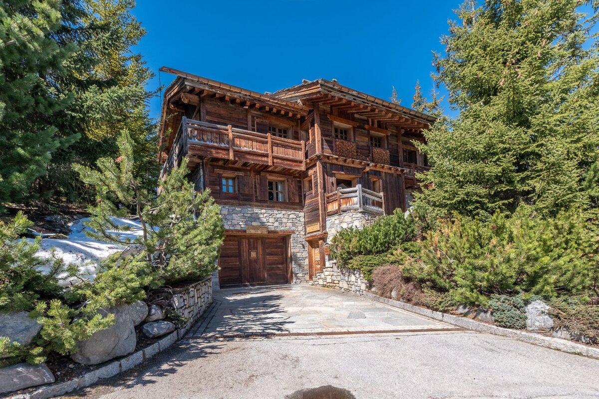 A rustic wooden alpine chalet with a stone basement and garage, nestled among pine trees under a clear blue sky.
