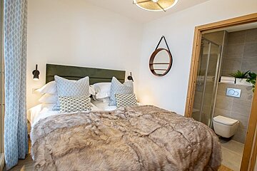 Cozy bedroom with a fluffy fur blanket, green headboard, patterned pillows, round wall mirror, and an adjacent en-suite bathroom.