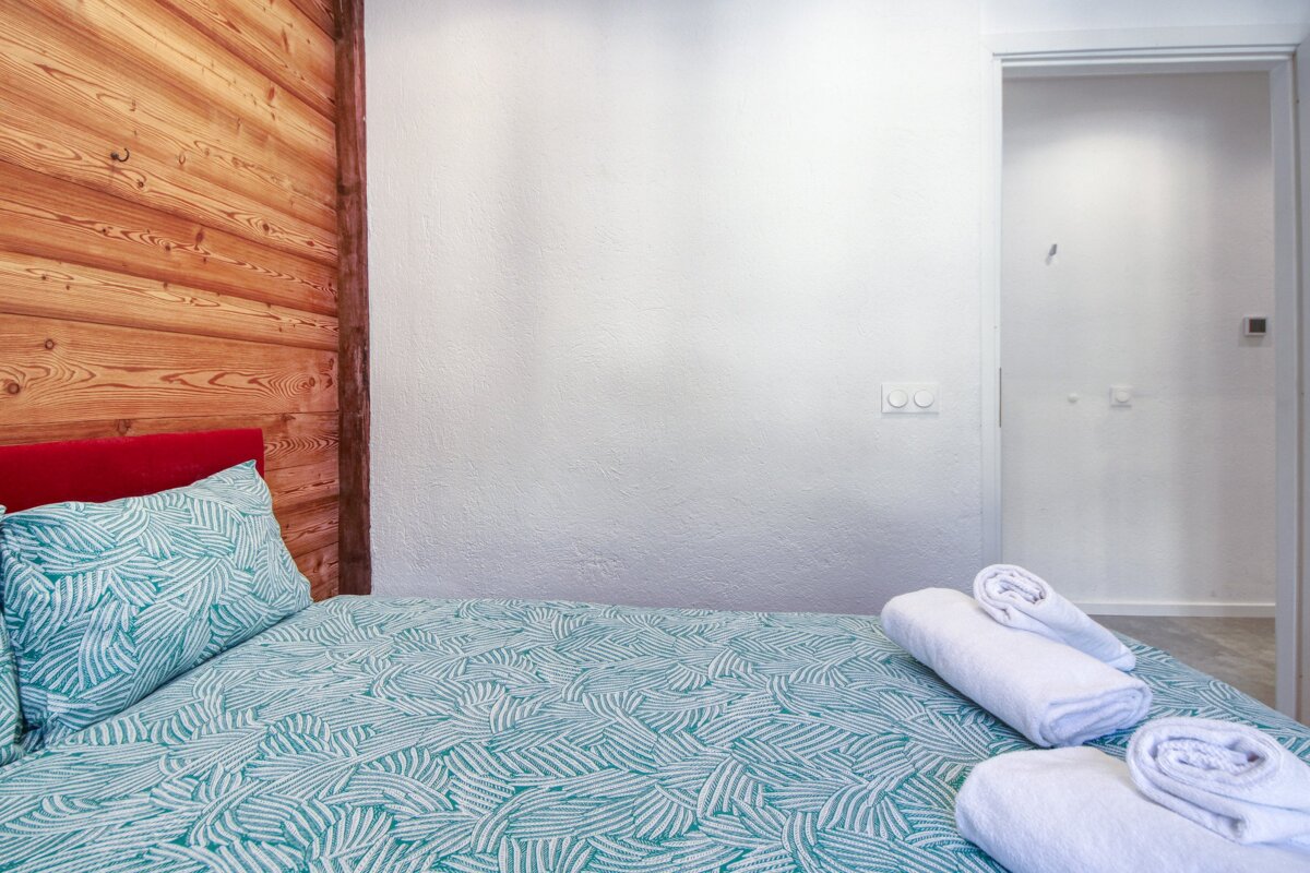 A cozy bedroom features a rustic wooden accent wall next to a plain white wall. The bed has a red headboard, teal patterned bedding, and fresh white towels.