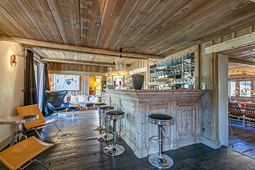 Rustic chalet interior featuring a wooden bar with chrome stools, wood-planked ceilings, and modern lounge chairs on dark floors.