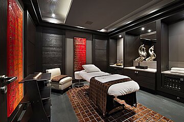 A sleek, dark spa room featuring a massage bed, vibrant red abstract art, elegant modern decor, a patterned brown rug, and soft lighting.