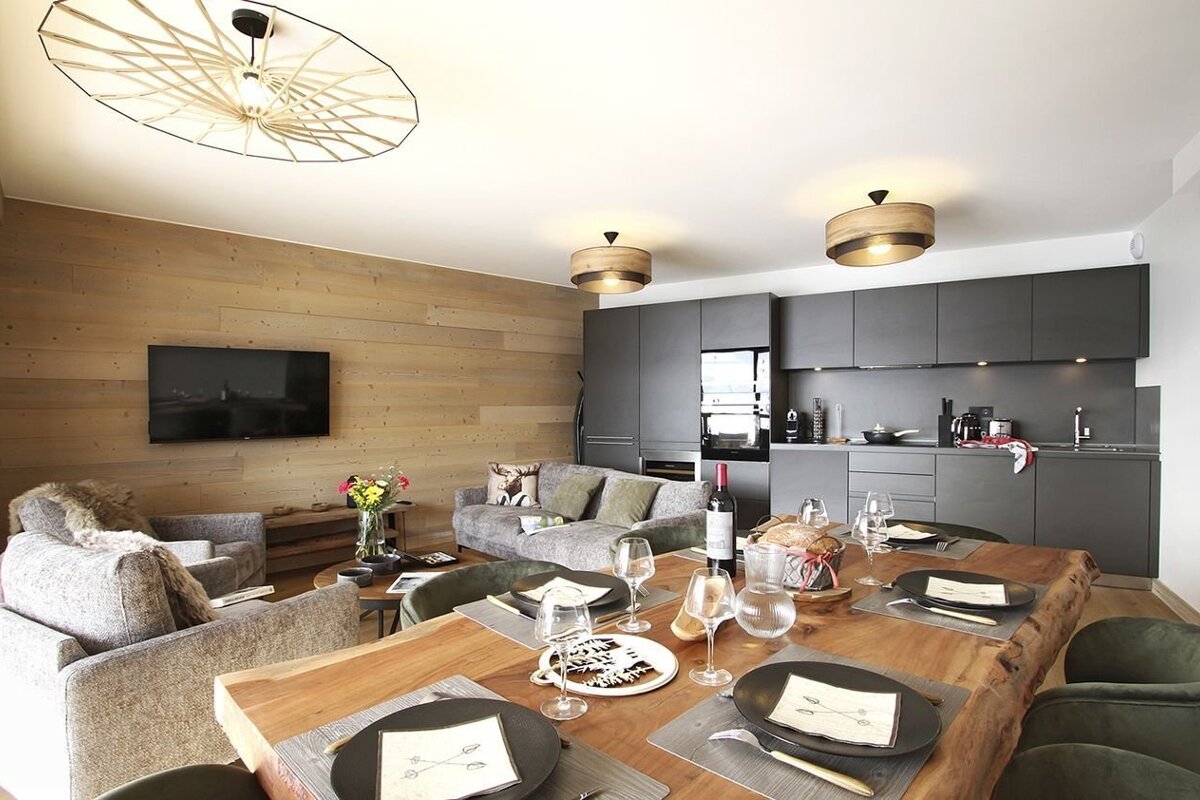 Cozy open-plan living and dining with a wooden wall and TV, adjacent to a modern dark grey kitchen. A rustic table is set for a meal.