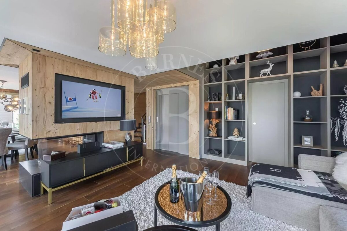 A luxurious, modern living room featuring wood paneling, a large TV displaying skiing, decorative shelving, a plush rug, and champagne. An elevator is visible.
