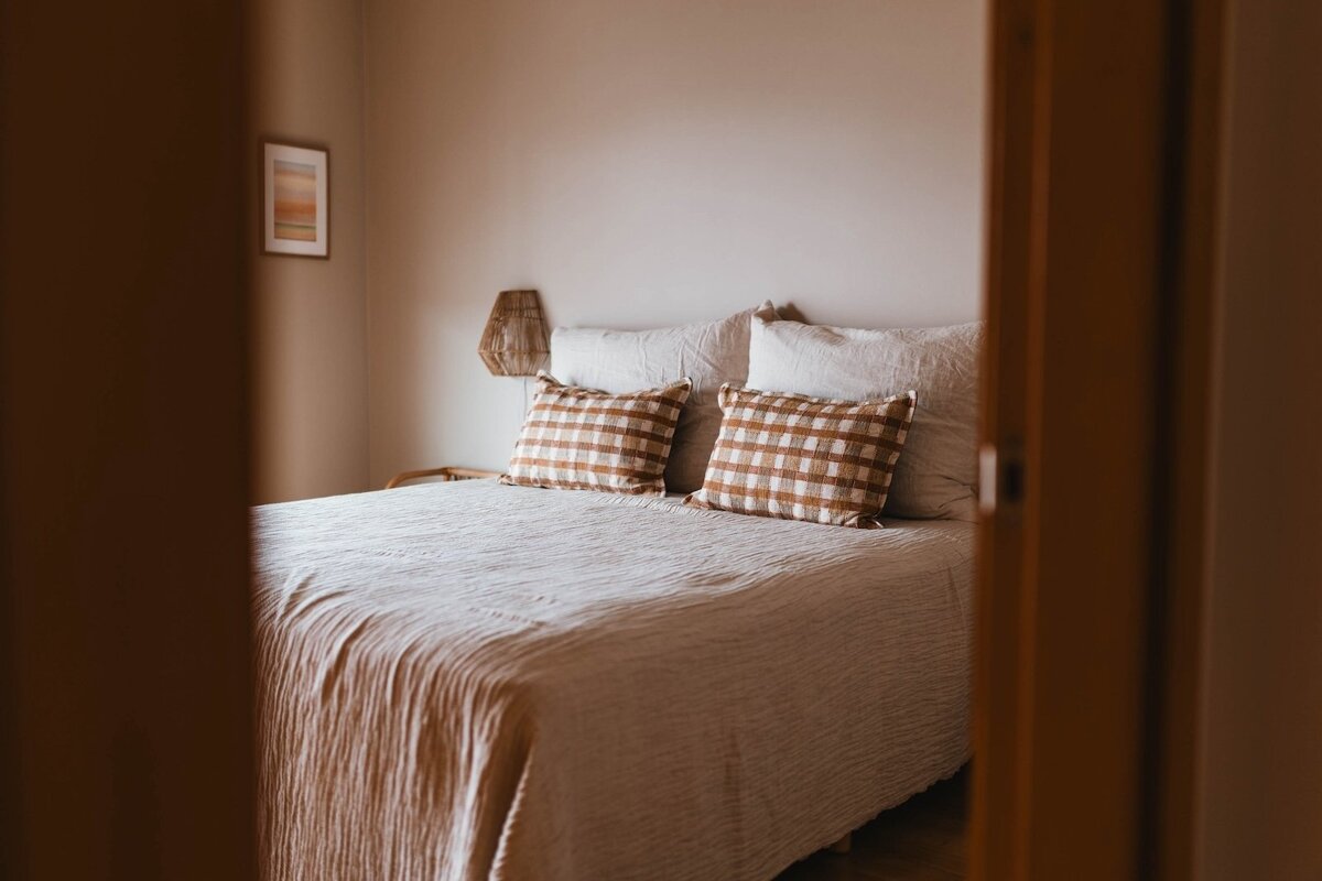 A cozy, neutral-toned bedroom with a bed featuring light linen bedding and two brown plaid pillows, framed by a wooden doorway.