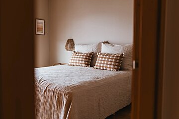 A cozy, neutral-toned bedroom with a bed featuring light linen bedding and two brown plaid pillows, framed by a wooden doorway.
