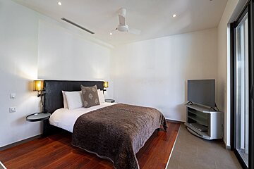 A minimalist bedroom featuring a bed with a brown cover, a dark headboard, wood flooring, a ceiling fan, and a TV.