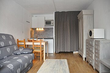 A small studio apartment featuring a grey patterned sofa, wooden dining set, kitchenette, and wood floors. Grey furniture like a wardrobe and drawers are also visible.
