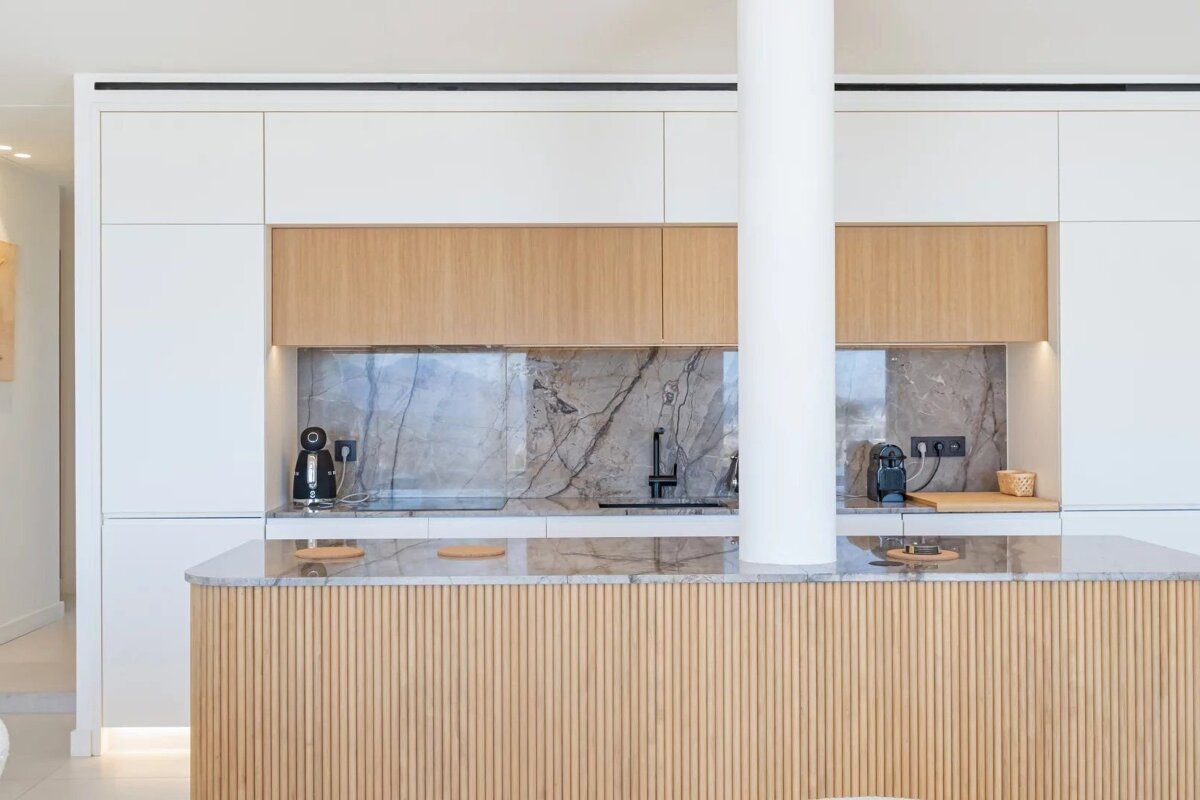 Modern minimalist kitchen featuring white cabinetry, light wood accents, a grey marble backsplash, and a slatted wooden island.