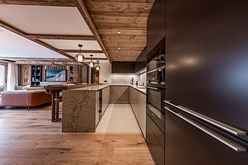 A spacious, modern open-plan kitchen and living room featuring rustic wood beams, sleek dark cabinetry, and light wood flooring.