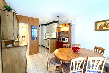 A rustic dining room features a round wooden table, plaid chairs, wood paneling, and a view into the kitchen.
