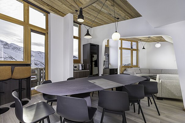 Sleek, modern dining/living room in a ski chalet with wooden ceilings, black dining set, and large windows revealing snowy mountain views.
