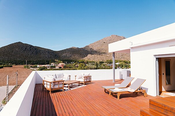 Sunny rooftop terrace with wood deck, lounge furniture, and sunbeds, offering mountain views and a distant village under a clear sky.