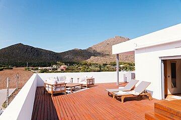 Sunny rooftop terrace with wood deck, lounge furniture, and sunbeds, offering mountain views and a distant village under a clear sky.