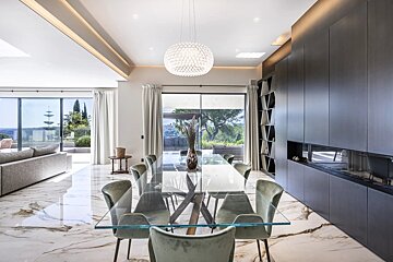 A modern dining room with a glass table, green chairs, marble floors, a large black cabinet, and a crystal chandelier.