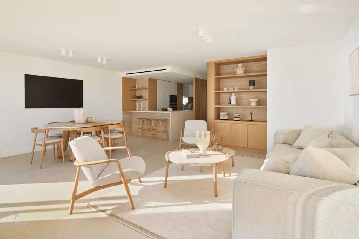 A bright, minimalist open-plan living area and kitchen with light wood furniture, cream textiles, and white walls, bathed in natural light.