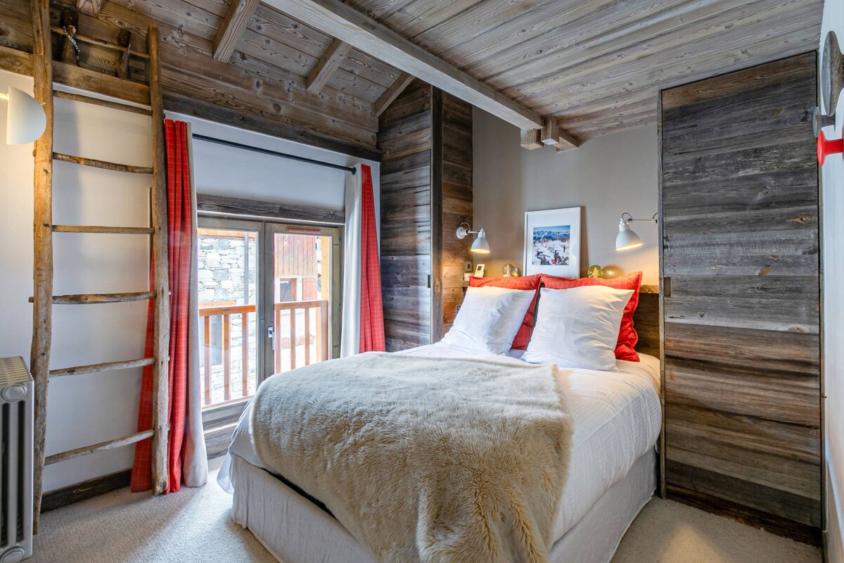 Rustic chalet bedroom with wooden walls and ceiling, a bed with red pillows and fur throw, and a balcony view. A ladder adds charm.