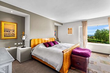 An inviting bedroom features a polished wooden bed with purple accents, an artwork, and a large window revealing a beautiful green landscape view.