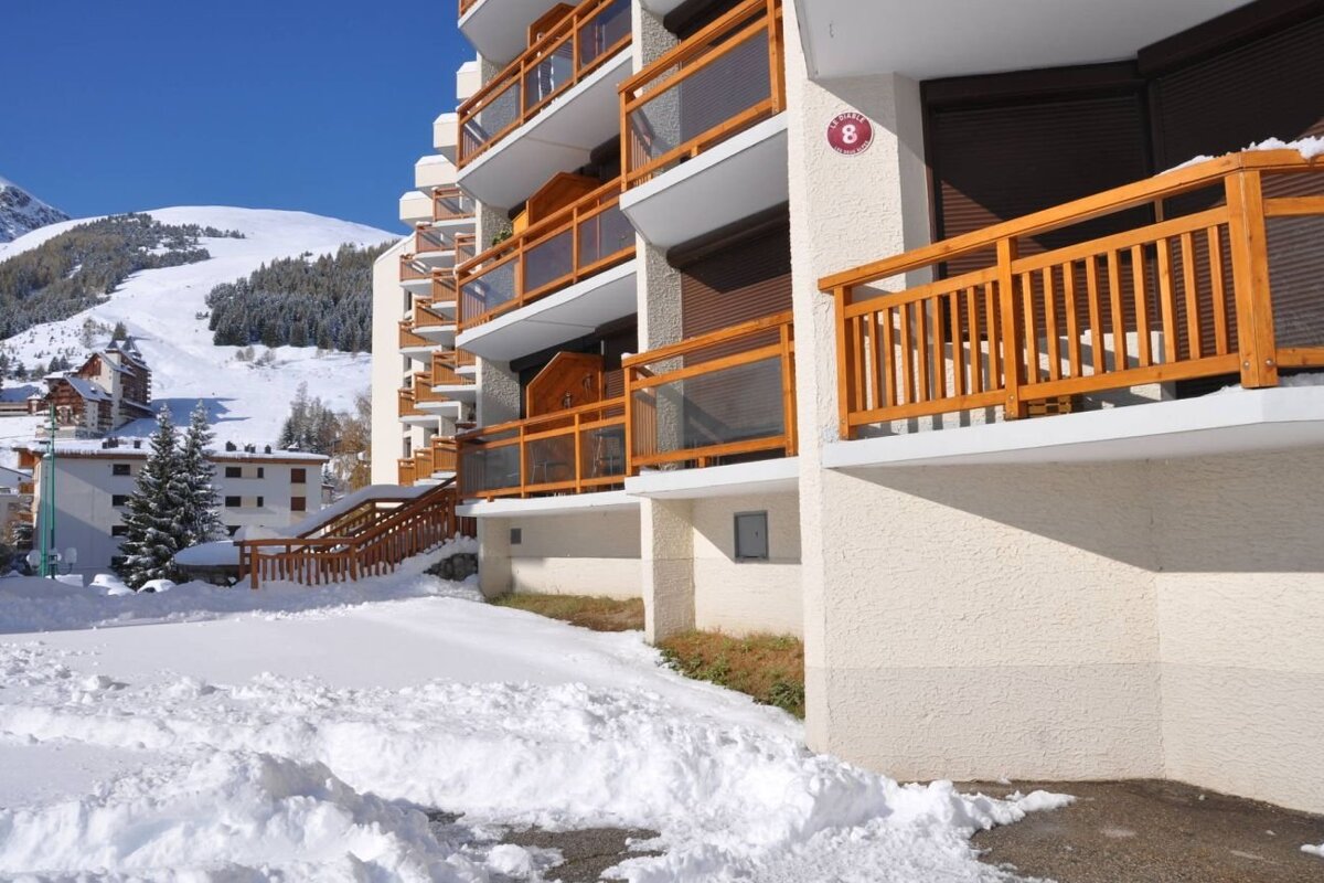 Alpine resort building with wooden balconies in a snowy landscape, with ski slopes and mountains in the background.