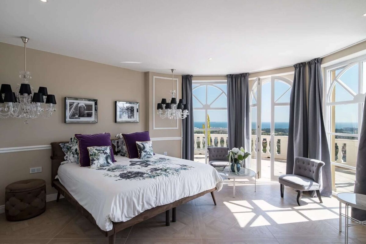 Elegant bedroom with a large bed, crystal chandeliers, and arched balcony doors revealing a stunning ocean view, bathed in natural light.