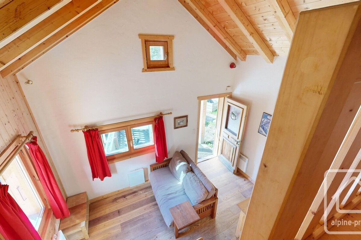 High-angle view of a cozy room with wooden floors, beams, and window frames. Features white walls, red curtains, a sofa bed, and an open door to outside.