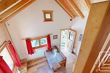 High-angle view of a cozy room with wooden floors, beams, and window frames. Features white walls, red curtains, a sofa bed, and an open door to outside.