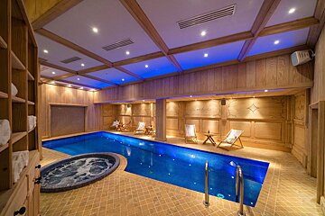 Luxurious indoor pool and bubbling jacuzzi in a wood-paneled room with serene blue ceiling lights and deck chairs.