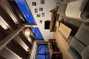 A modern, well-lit living room with a high wooden beamed ceiling, large windows showing blue, a gray sofa, wooden dining table, TV, and framed art.