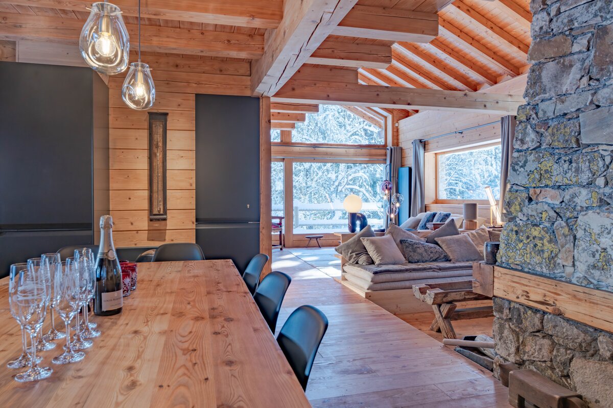 Cozy alpine chalet interior featuring a wooden dining table, stone fireplace, and large windows looking out onto a snowy winter landscape.