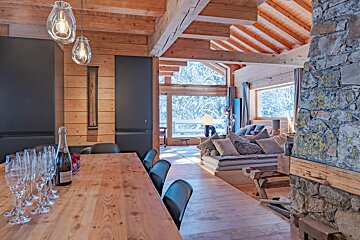 Rustic wooden chalet with a champagne-set dining table and a cozy living area. Large windows frame a beautiful snowy winter landscape outside.