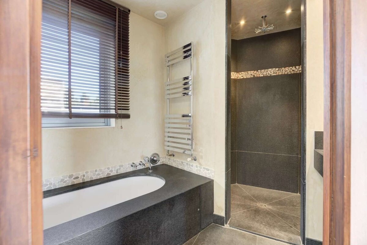 Modern bathroom with a dark stone bathtub, window with wooden blinds, towel warmer, and a walk-in shower with dark tiles and pebble accent.