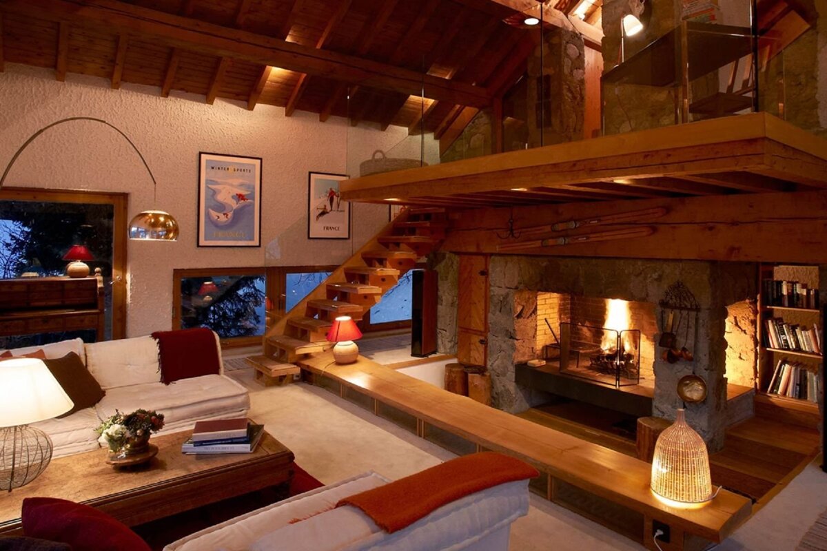 A cozy, multi-level ski lodge living room with a stone fireplace, exposed wooden beams, floating stairs, and a mezzanine, illuminated by warm lighting.